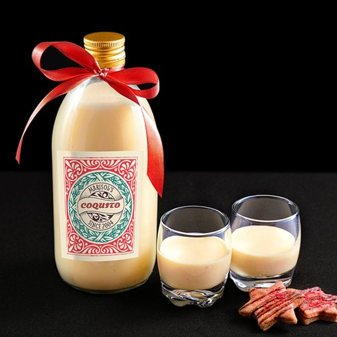 A clear glass bottle full of Coquito with a custom label and tied with a red ribbon for holiday gift giving.